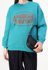 Teal sweatshirt med en stor brun "AMERICAN VINTAGE" logotyp, tillsammans med en liten svart läderhandväska. Bär mörka pinstripade byxor.