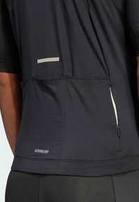 Black athletic shirt with a textured finish, featuring a back zipper pocket and a reflective stripe detail. Logo displayed at the bottom.