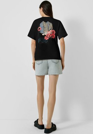 Kobieta w czarnej koszulce z kolorowymi paskami i grafiką homara na plecach, w połączeniu z jasnymi szortami dżinsowymi i czarnymi sandałami na platformie.