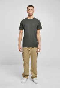 T-shirt grigio scuro a maniche corte, pantaloni cargo beige con tasche laterali e sneakers bianche; in piedi contro uno sfondo grigio chiaro.