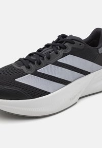 Schwarzer Sportschuh mit strukturiertem Mesh-Obermaterial, ausgestattet mit drei reflektierenden silbernen Streifen. Er hat eine dicke weiße Sohle mit geriffelten Akzenten.