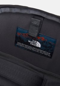 Schwarzes Stoffinnere einer Tasche, mit einem blauen und orangefarbenen Etikett, das das "The North Face"-Logo und Informationen über recycelte Materialien zeigt. Reißverschlussdetails sichtbar.