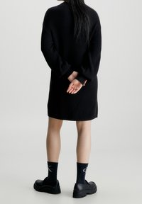 Robe pull noire à côtes avec manches longues, présentant une coupe décontractée et un ourlet au genou. Portée avec des chaussures noires épaisses et des chaussettes à motifs.