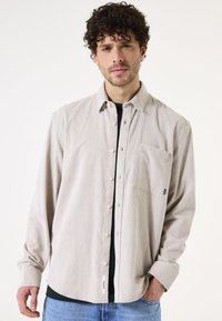 Lichtbeige overhemd met knopen en een zak, van een textuurstof, met lange mouwen en een gebogen zoom, gedragen over een zwart shirt.