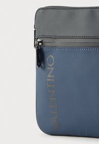 Μπλε υφασμάτινη τσάντα με υφή στην επιφάνεια, με μαύρο επάνω μέρος, ασημένια μεταλλικά στοιχεία και ανάγλυφο λογότυπο στο πλάι.