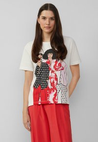 T-shirt en coton blanc avec une impression graphique de trois femmes vêtues de tenues vives. Les couleurs notables incluent le noir, le rouge et le rose avec des pois.