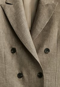 Blazer en lin marron présentant un design croisé, des revers crantés et des boutons noirs avec une texture lisse et une légère brillance.