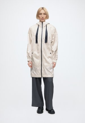 Femme aux cheveux blonds courts portant une longue veste à capuche beige clair avec des cordons noirs, un pantalon large foncé et des chaussures noires, debout face à l’avant.