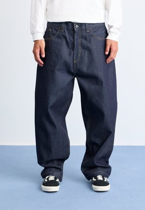 Mørkeblå denimjeans med en løs pasform, der har gule syninger, fem-lomme-design og knaplukning. Båret med sort og hvide sneakers.
