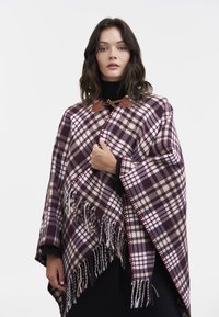 Poncho de cuadros en burdeos y crema, hecho de tela suave. Presenta bordes con flecos y cierres de toggles marrones en el cuello.