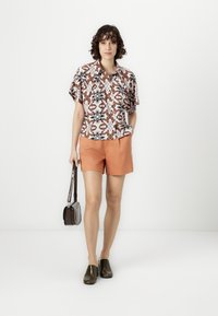 Short-sleeve floral shirt with orange shorts, black slip-on shoes, and a small brown handbag. Bright colors and patterned fabric details.