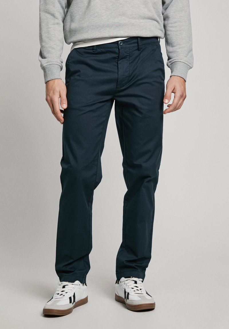 Homme portant un pantalon bleu foncé, des baskets blanches avec des rayures noires et un sweat-shirt gris clair, debout devant un fond uni.