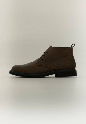 Bottine en daim marron avec semelle noire, trois œillets à lacets et languette au talon, présentée en profil sur fond neutre.