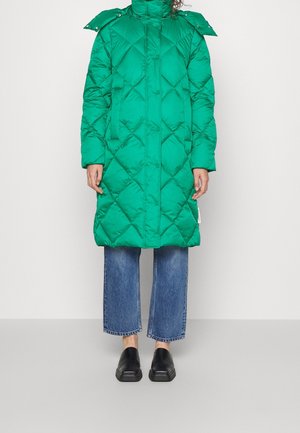 Green quilted long puffer coat worn with blue cropped jeans and black slip-on shoes, standing against a white background.
