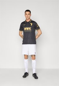 Crni nogometni dres s zlatnim akcentima, s uzorkom. Uparen s bijelim šortsevima, crnim kopačkama i bijelim čarapama.