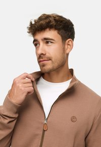 Brown ribbed zip-up jacket with a collar, featuring a leather tag logo and a white t-shirt underneath. Smooth texture, casual design.