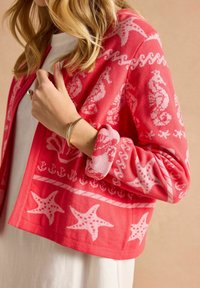 Korálový pletený cardigan s vzorem mořského koníka a hvězdice. Texturovaná tkanina s volným střihem a ohrnutými manžetami.