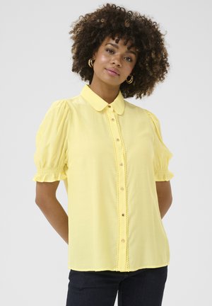 Camicia gialla con bottoni, maniche corte a sbuffo, caratterizzata da un sottile bordo testurizzato lungo la parte anteriore e bottoni naturali. Tessuto liscio.