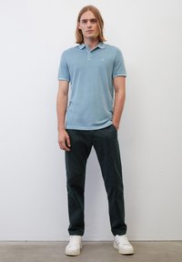 Helles blaues Poloshirt mit Kragen, kombiniert mit dunkelgrünen Hosen und weißen Turnschuhen. Glattes Material mit dezentem Logo-Detail auf der Brust.