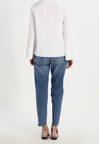 Blouse blanche à manches longues avec des poignets évasés, associée à un jean en denim bleu. Le jean présente un ourlet effiloché et une coupe décontractée. Des sandales noires complètent la tenue.
