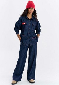 Zestaw z kurtką i spodniami z denimu w ciemnym niebieskim kolorze z czerwonymi akcentami. Kurtka posiada duże kieszenie; spodnie mają luźny krój. Model nosi czerwoną czapkę typu beanie.