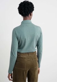 Maglia a maniche lunghe color turchese con collo alto realizzata in tessuto morbido, abbinata a pantaloni marroni in velluto a coste svasati che mostrano una texture a coste verticali.
