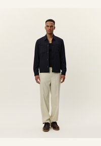 Jachetă cu dungi navy, cu guler, nasturi frontal și două buzunare pe piept, asortată cu pantaloni bej și încălțăminte închise la culoare. Design simplu, modern.