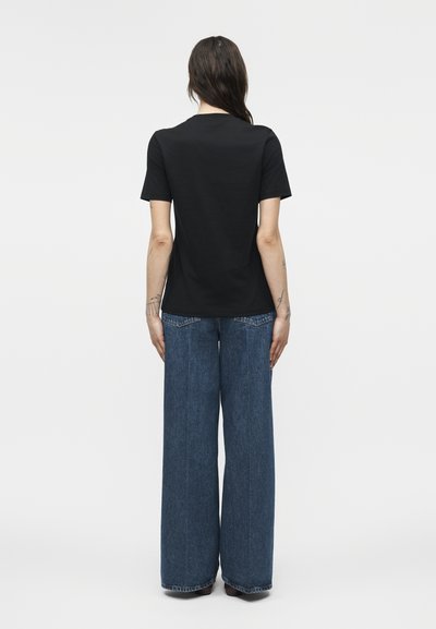 Γυναίκα που στέκεται με την πλάτη γυρισμένη, φοράει απλό μαύρο κοντομάνικο μπλουζάκι και φαρδιά μπλε τζιν παντελόνια μπροστά σε λευκό φόντο.