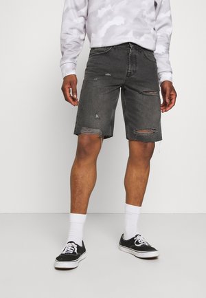 Homme portant un short en jean noir déchiré, des chaussettes blanches montantes, des baskets noires et blanches et un haut à manches longues de couleur claire.