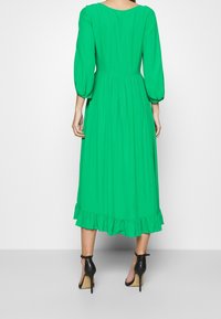 Vihreä, polvipituinen mekko, jossa on pyöreä pääntie, puffat pitkät hihat, kootut vyötärö ja röyhelöinen helma. Päällä mustat korkokengät.