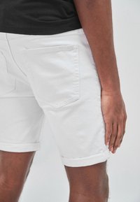 Shorts de algodón blancos con puños enrollados, que cuentan con un bolsillo trasero y una textura suave. La prenda está usada y parcialmente visible en una persona.