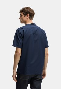 Camicia blu navy a maniche corte, texture liscia, colletto a punta, cuciture in contrasto e vestibilità rilassata. La vista posteriore mostra dettagli di design minimalisti.