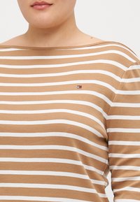 Braun-weißer gestreifter Langarm-Top mit weitem Halsausschnitt; verfügt über ein kleines Logo auf der linken Brust. Hergestellt aus weichem, leichtem Stoff.
