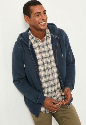 Navy zip-up hoodie over a beige checkered button-up shirt. The hoodie features drawstrings and a textured finish. Olive green pants complete the outfit.