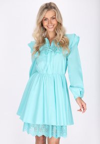 Rochie turcoaz cu mâneci lungi, umeri fluturați, talie ajustabilă cu șnur și tiv din dantelă. Material neted cu accente de cusături decorative.