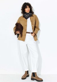 Femme aux cheveux bouclés portant une veste beige, un pantalon blanc, une écharpe noire à motifs, des bottes en imprimé léopard et tenant un sac en cuir frangé marron.