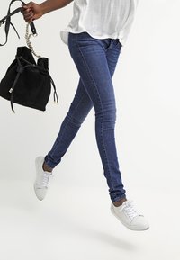 Jeans de mezclilla azul skinny, diseño ajustado, cintura de altura media, combinados con zapatillas blancas y un bolso de cubo negro con un asa de cadena.
