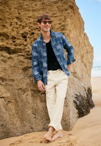 Giovane uomo con occhiali da sole che indossa una giacca blu a toppe, una maglietta nera e pantaloni beige, appoggiato a piedi nudi contro una grande roccia sulla spiaggia.