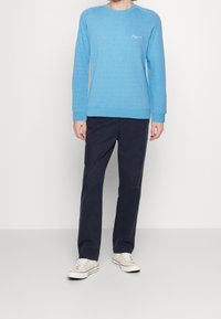 Sudadera de textura azul claro con manga larga y puños acanalados, combinada con pantalones de color azul marino y zapatillas blancas. Detalle de logo sutil en el pecho.