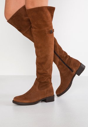 Personne portant des bottes en daim marron montantes avec fermetures éclair latérales, petit détail de boucle, talons bas, marchant sur un sol clair.
