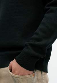 Άτομο που φοράει ένα σκούρο πλεκτό πουλόβερ με το ένα χέρι χαλαρά χωμένο στην μπροστινή τσέπη από μπεζ τζιν παντελόνι.