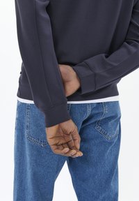 Felpa navy con maniche lunghe e colletto a girocollo, abbinata a jeans in denim azzurro. Mano posizionata dietro la schiena.