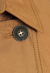 Detailní záběr hnědé látky s detailním šitím a velkým černým knoflíkem se čtyřmi dírkami přišitém blízko překládaného okraje.