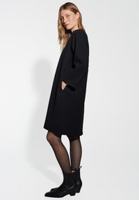 Zwarte wollen jurk met een losse fit, driekwart mouwen, zijzakken en een lengte tot de knie. Draagstijl met enkellaarzen en doorschijnende tights.