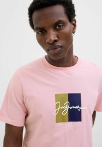 Lyserød bomulds t-shirt med grafisk design, der har rektangulære blokke i guld, blå og hvid tekst. Ribbet rund hals og korte ærmer.