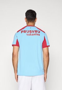 Vīrietis ar īsām piedurknēm gaiši zila un sarkana sporta krekla ar uzrakstu "PRIJSVRIJ vakanties" mugurā, stāv pret vienkāršu fona.