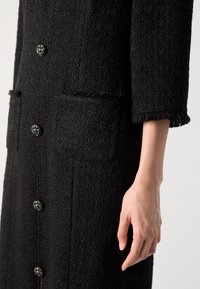 Cappotto nero con texture, dotato di chiusura a bottoni, due tasche frontali e orlo frangiato. I bottoni sono decorati con dettagli decorativi.