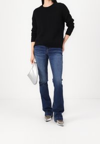 Person in einem schwarzen Pullover, blauen Jeans und silbernen, spitzen Schuhen, die eine kleine silberne Handtasche vor einem schlichten weißen Hintergrund hält.