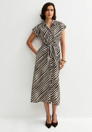 Kobieta stojąca, ubrana w czarno-białą sukienkę midi w ukośne paski, z paskiem wiązanym w talii i czarne czółenka na szpic.