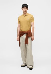 Μουσταρδί πόλο μπλούζα με κοντά μανίκια και ένα μικρό έμβλημα, συνδυασμένη με ανοιχτό μπεζ παντελόνι φαρδιάς γραμμής και πλεκτό πουλόβερ σε σκουριά χρώμα δεμένο γύρω από τη μέση.